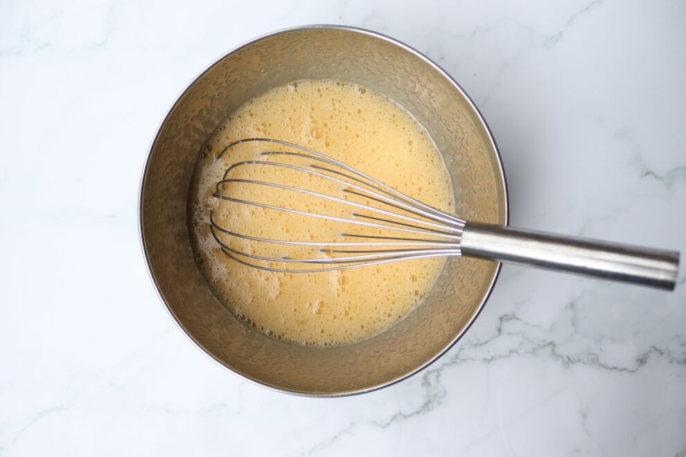 schaal met cake ingredienten, beslag met garde, beslag met melk, olie, vanillepasta, zout en eimengsel.